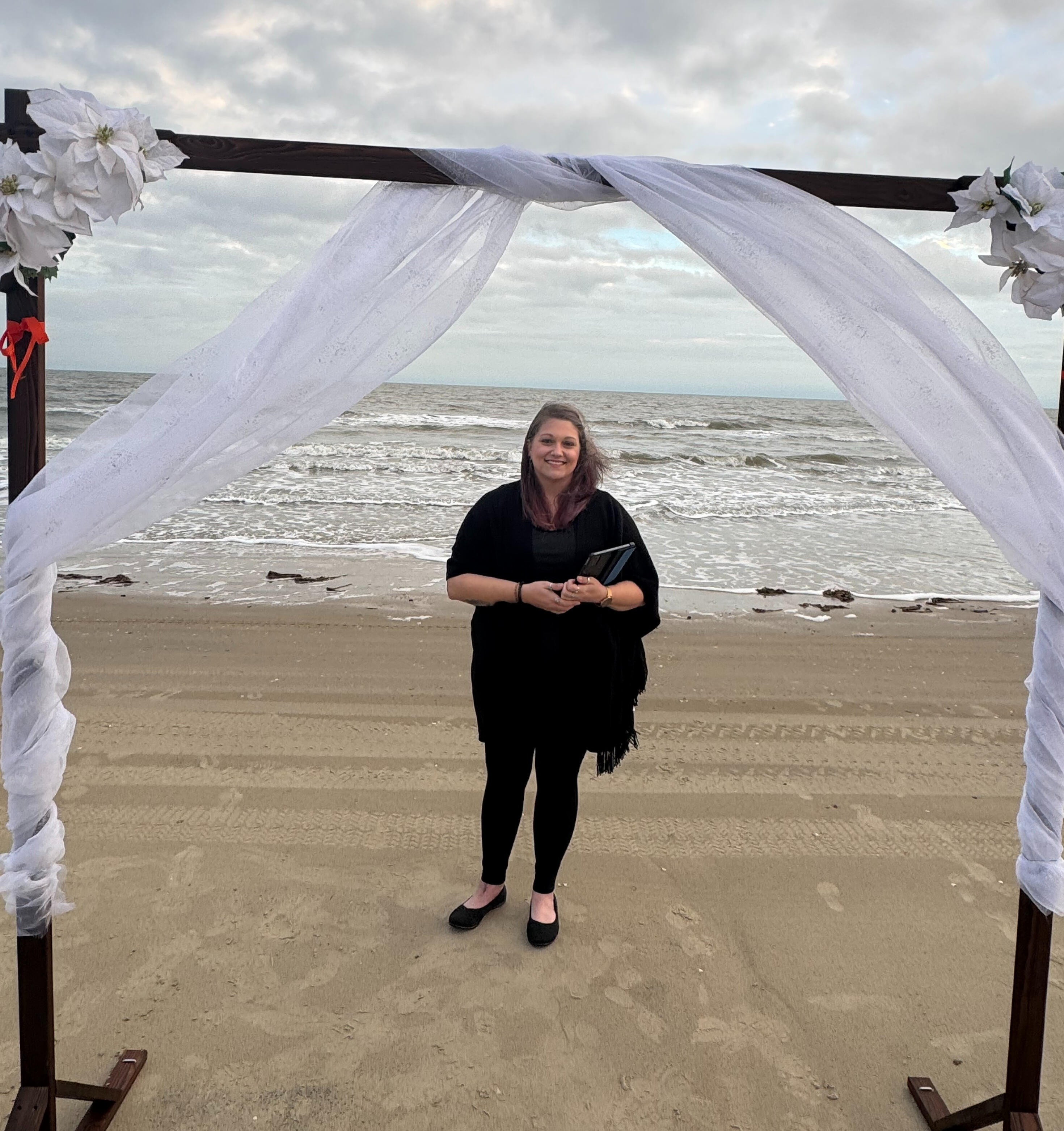 Rachel standing on the beach, smiling, wearing all black below an arch draped in flowy, white fabric with the ocean visible behind her