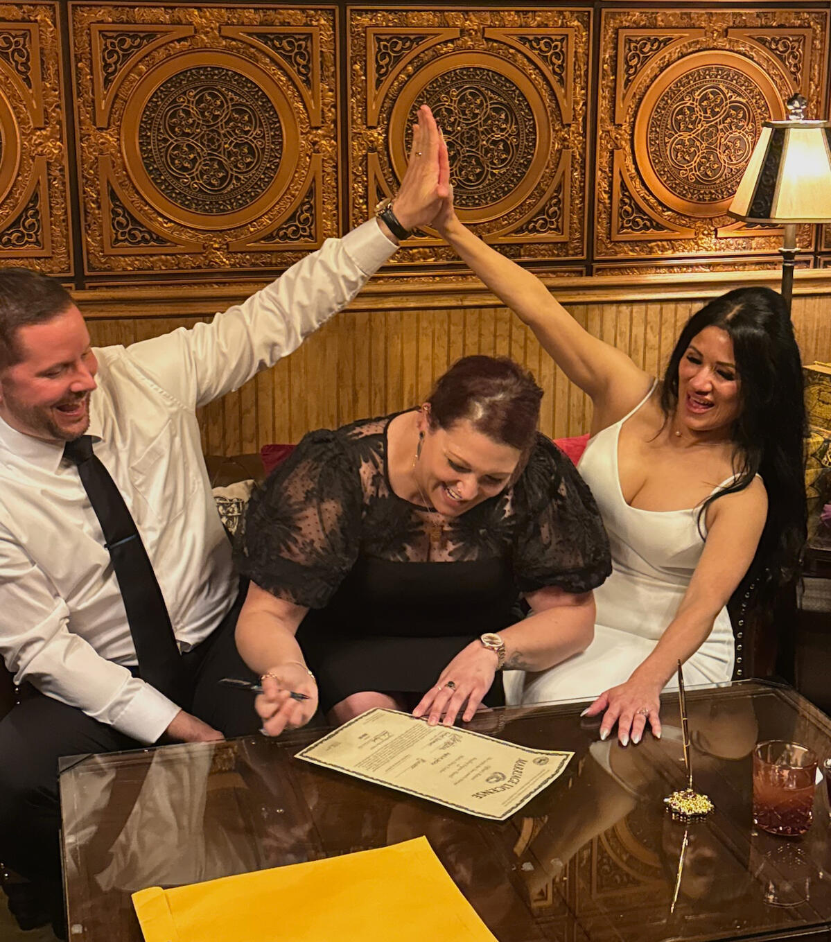 Couple sitting on a couch high-fiving above Rachel as she signs their marriage license
